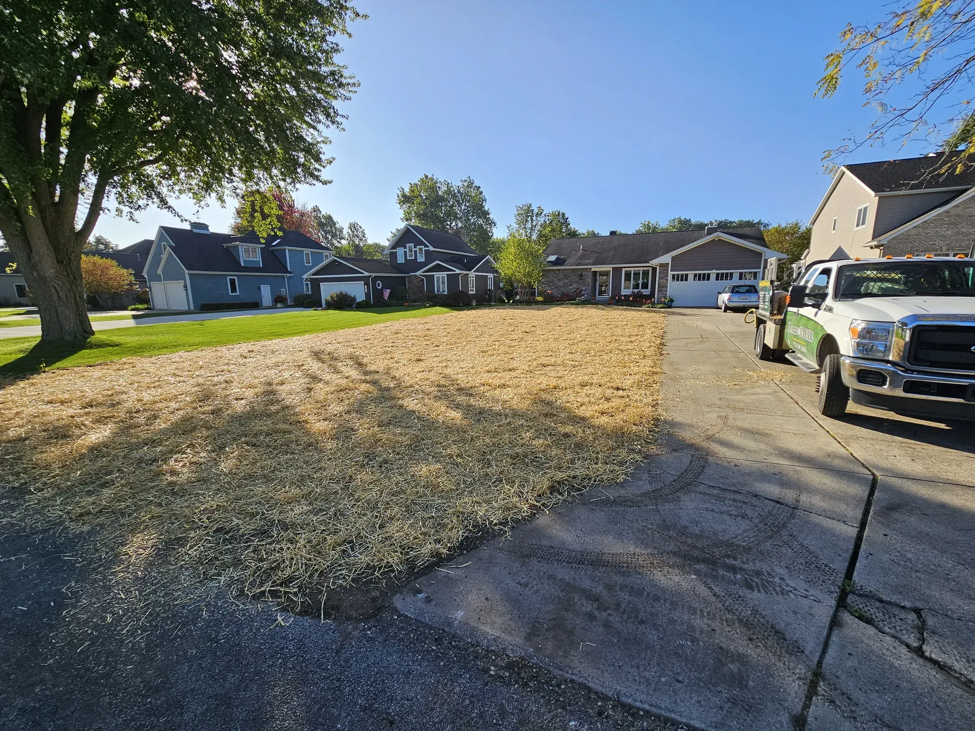 An irregular brown, dead patch of lawn beside healthy green turf, with a service truck parked in the driveway.