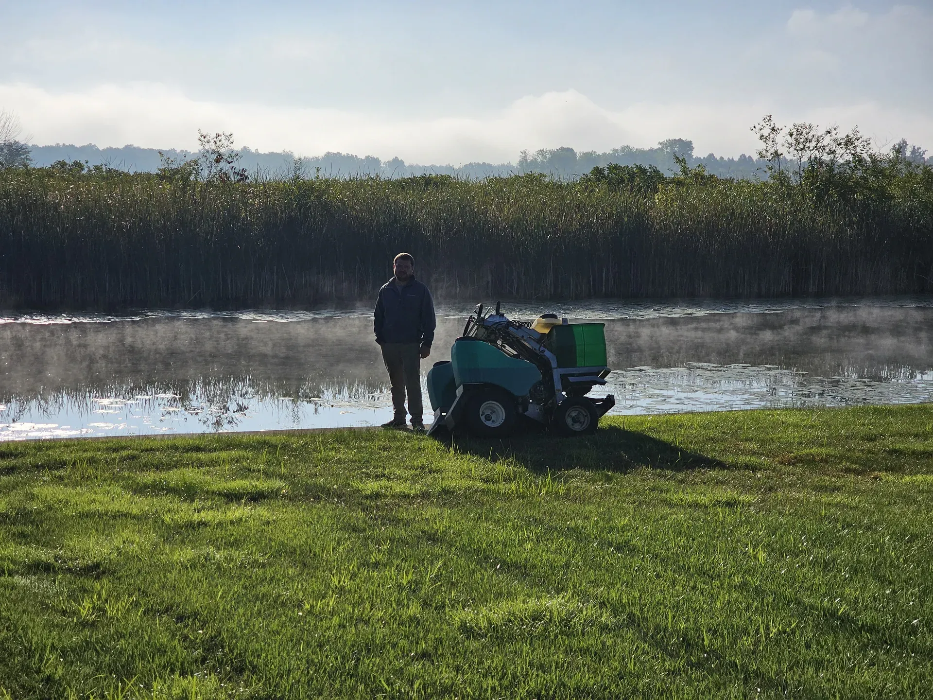 A technician stands beside a green ride-on core aerator at the foggy edge of a lawn along the water.