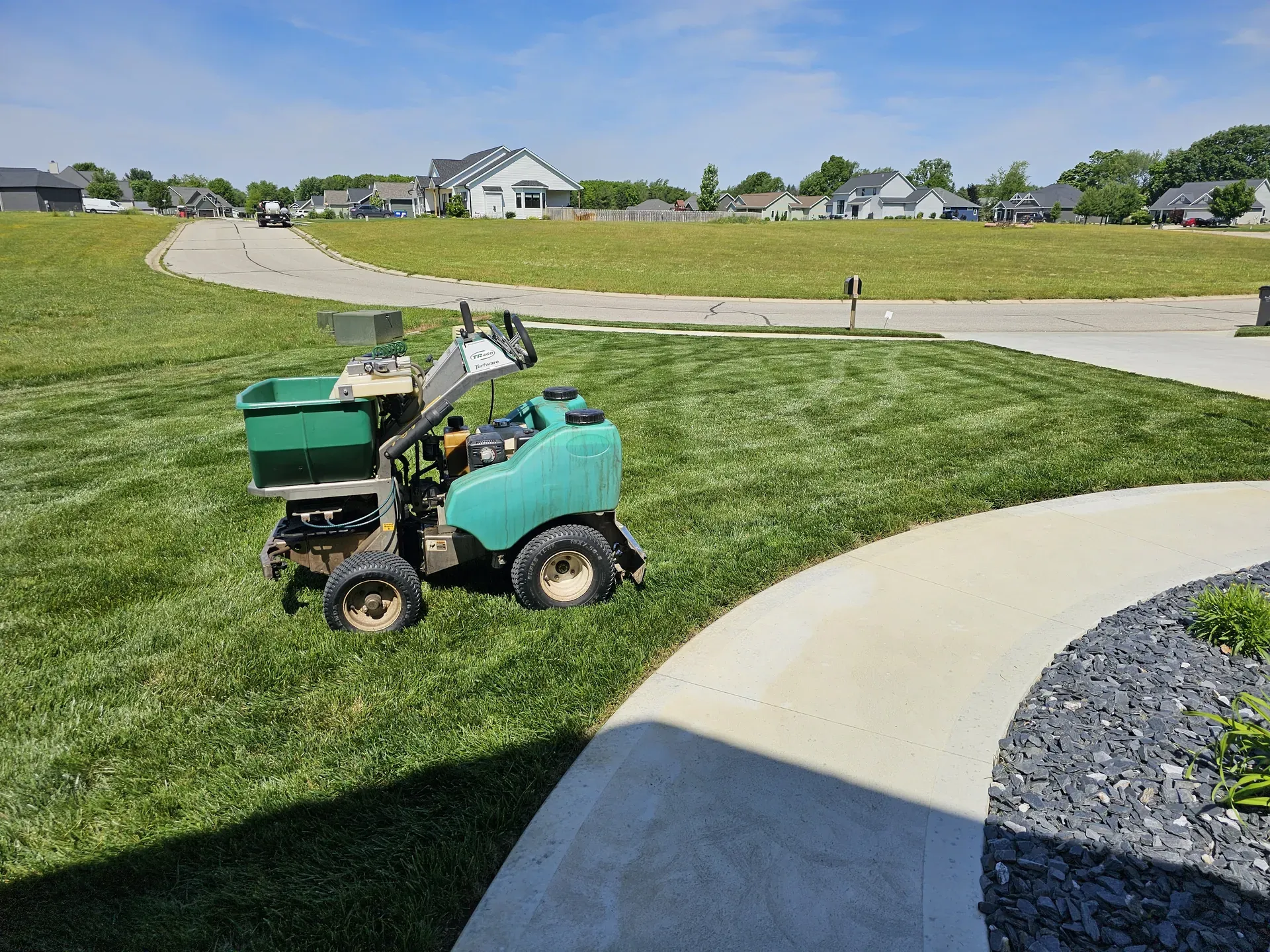 A ride-on granular spreader applying fertilizer on a striped lawn next to a sidewalk