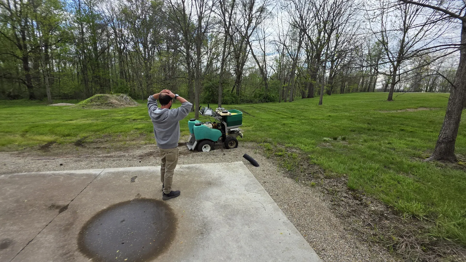 A technician sprays with a backpack sprayer near a wooded edge, with standing water on the pavement nearby.
